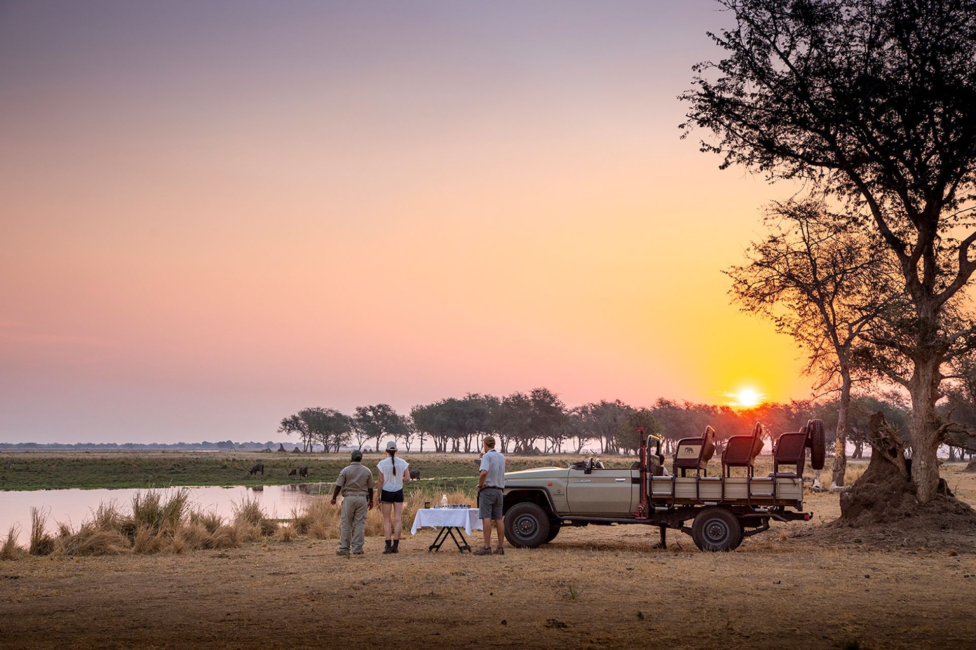 A safari vehicle parked at sunset by a riverbank with three people preparing a table for an outdoor meal.