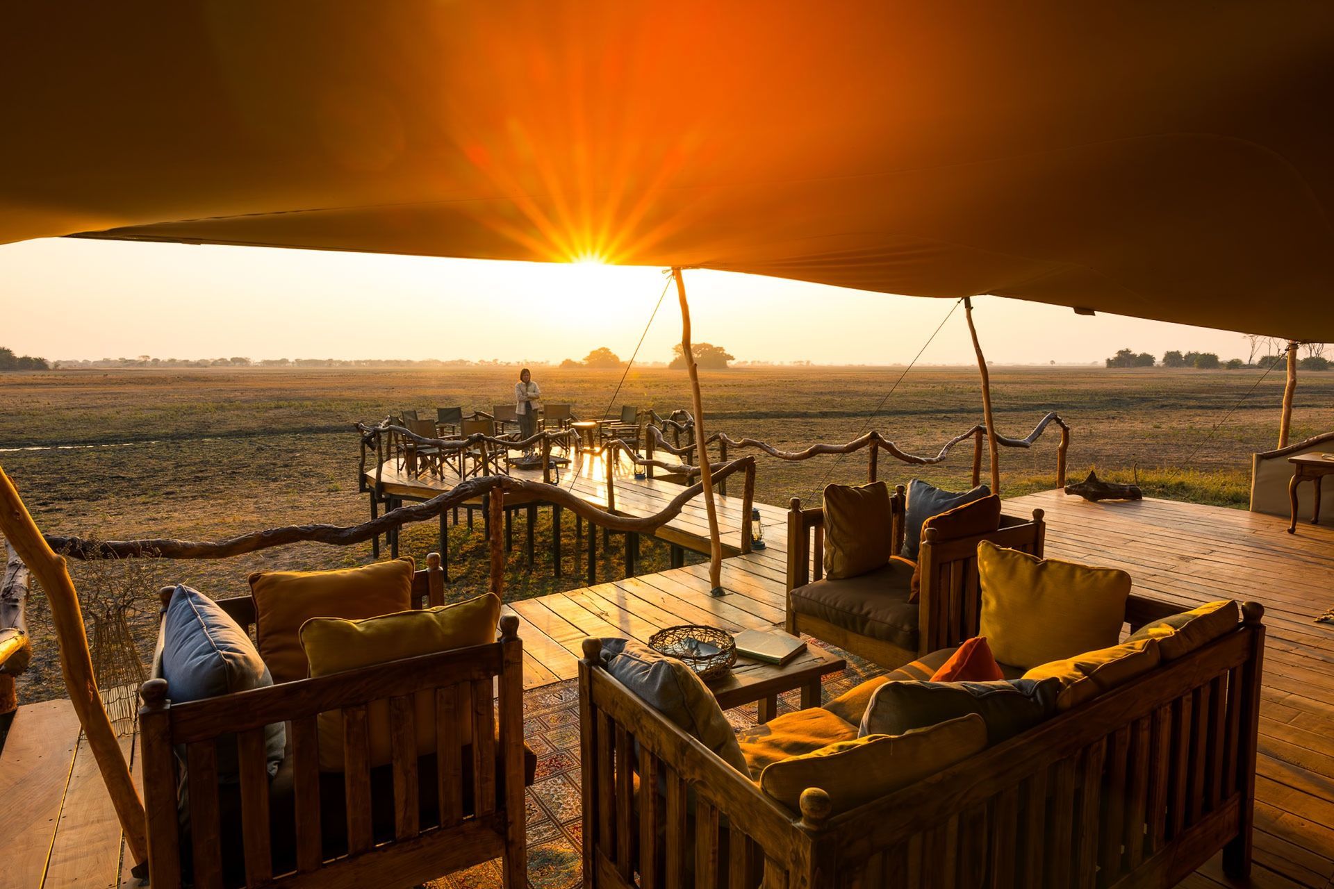 Sunset view from a safari tent deck with comfortable wooden lounge furniture looking out over an open, grassy savanna.