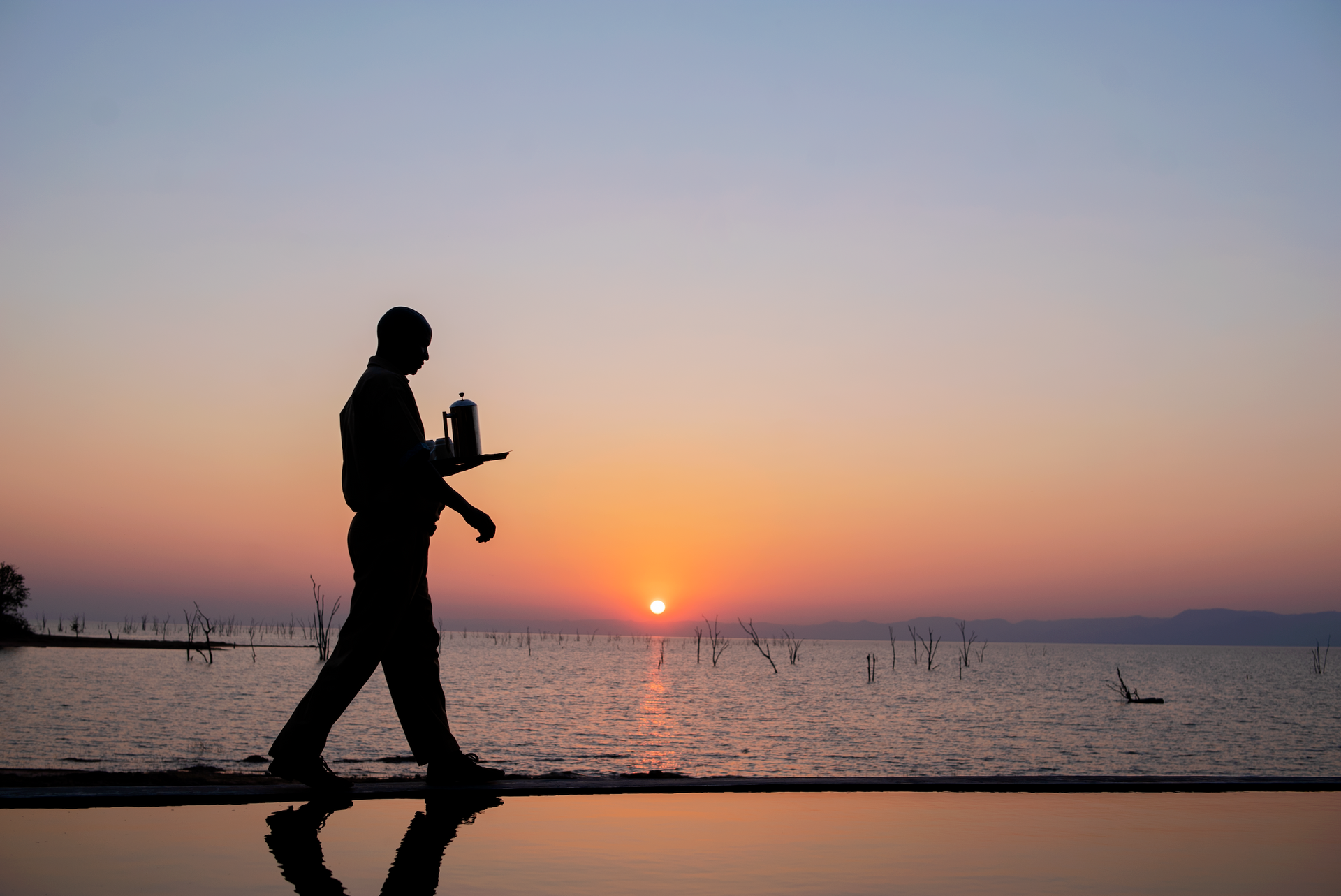 A silhouette of a person carrying a tray walks along a waterfront at sunrise.