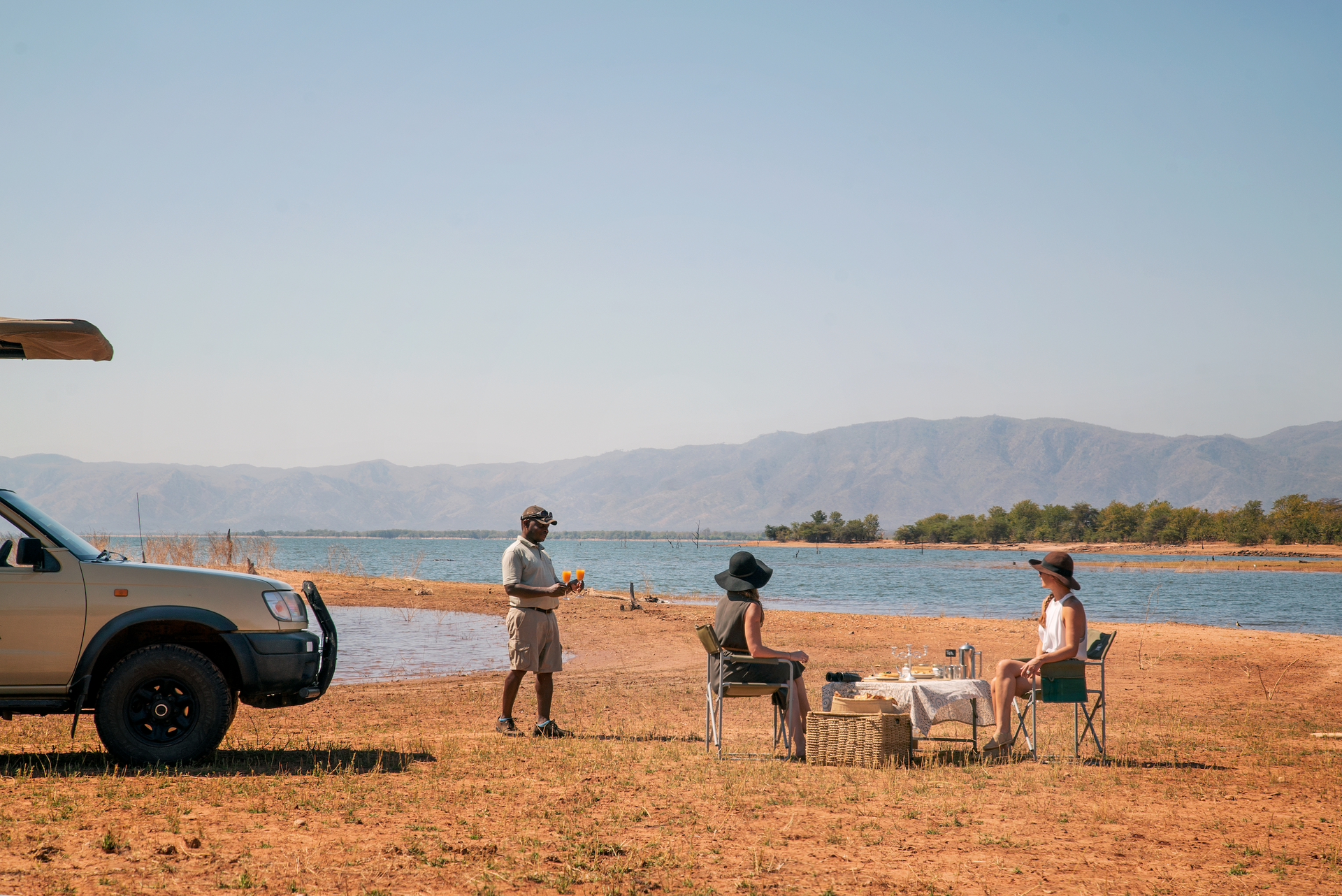 Two people sit at a table near a parked SUV by a lake, while a third person stands nearby in a sunny, arid landscape.