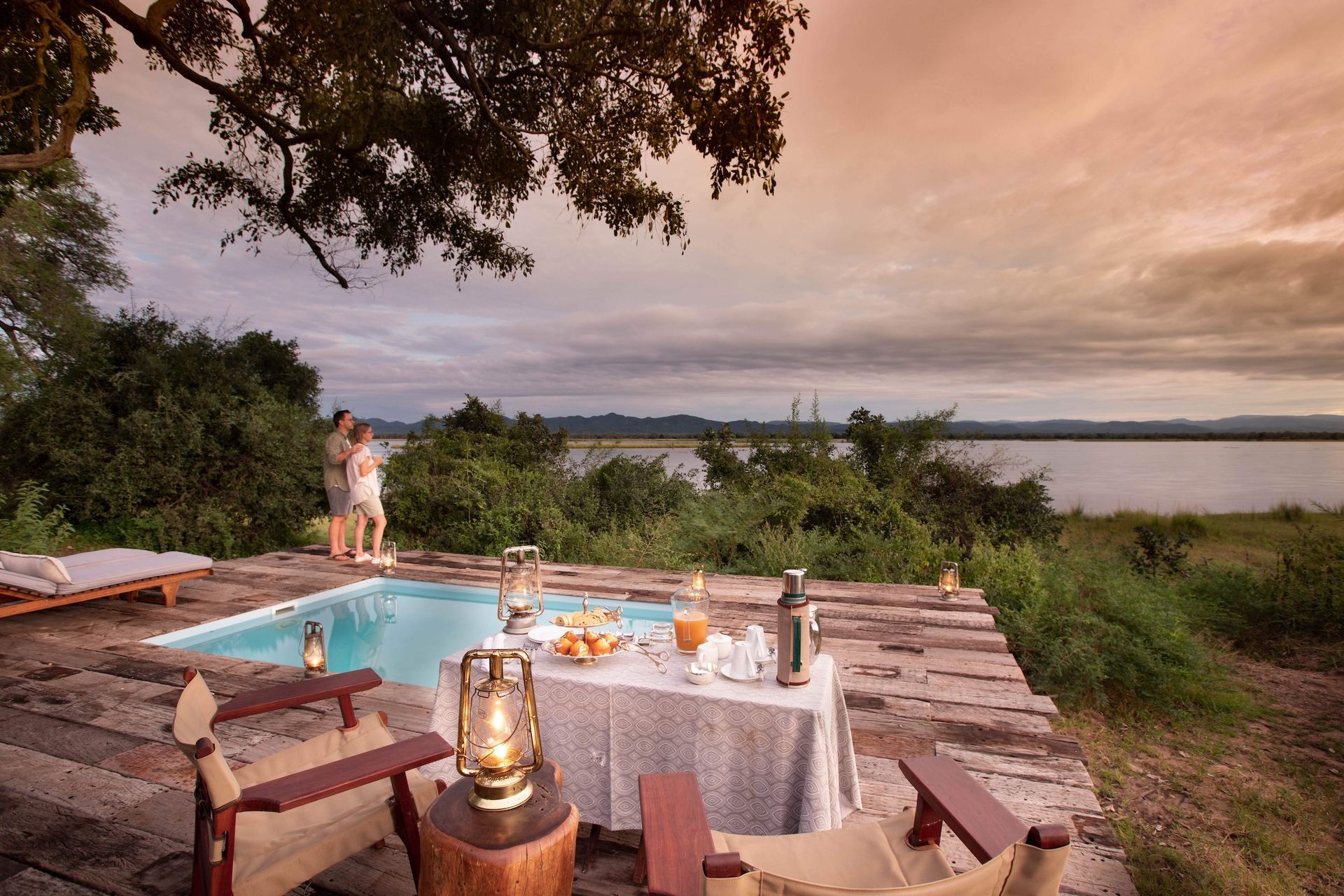 A couple stands by a pool and table at a sunset safari lodge overlooking a wide river.