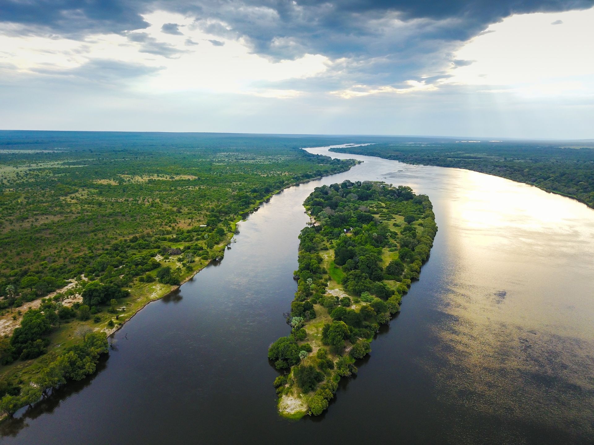 An aerial view of a long, tree-covered island in the middle of a wide, dark river surrounded by a lush green landscape.