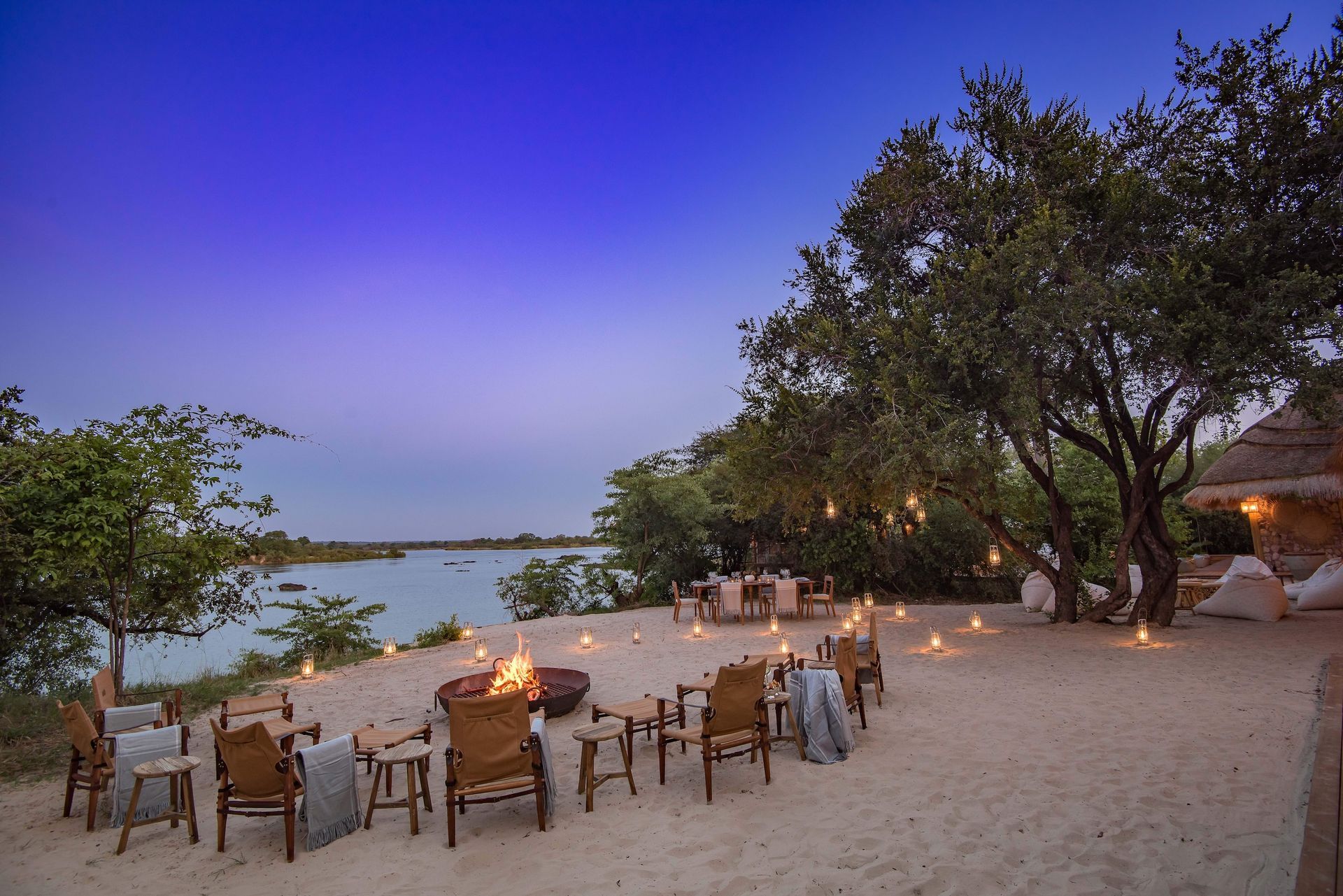 An outdoor lounge at twilight, with safari chairs arranged around a fire pit on a sandy bank overlooking a calm river.