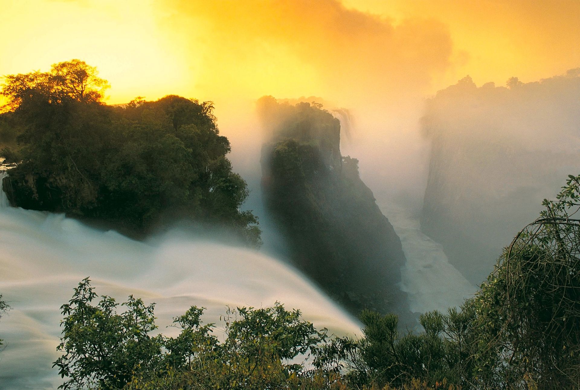 Golden light illuminates mist rising from the cascading waters and rocky cliffs of Victoria Falls at sunset.