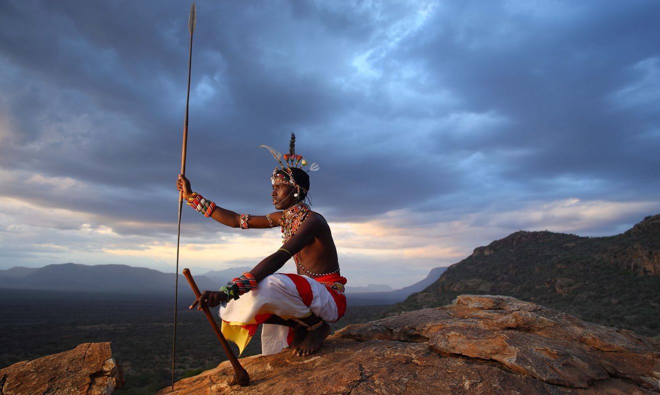 A person in traditional clothing holds a spear while crouching on a rocky mountain ridge against a cloudy sunset sky.