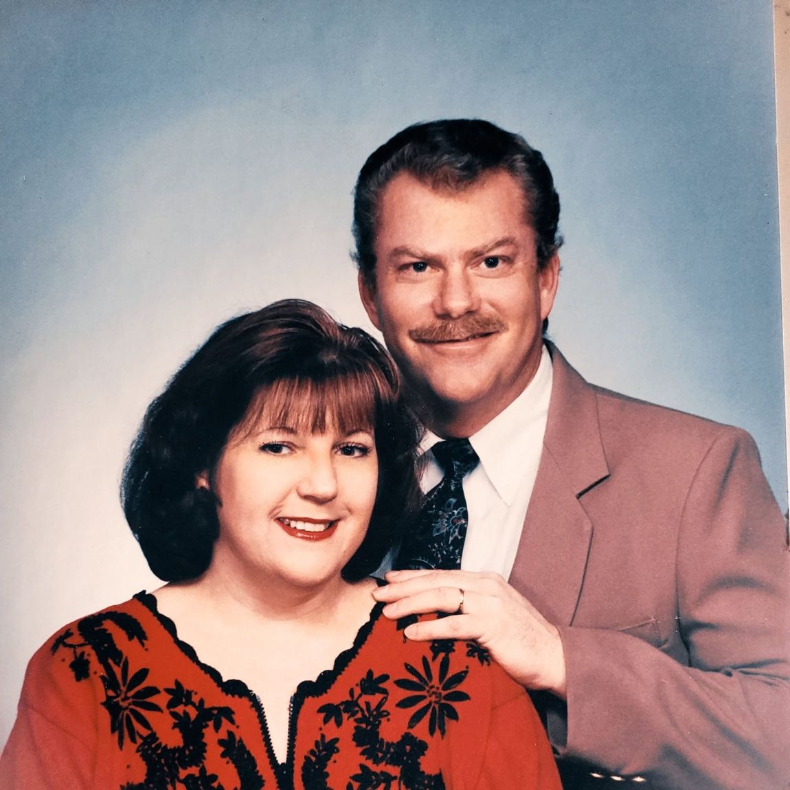 Smiling couple posing against a blue studio backdrop, the woman in a red floral top and the man in a tan suit.