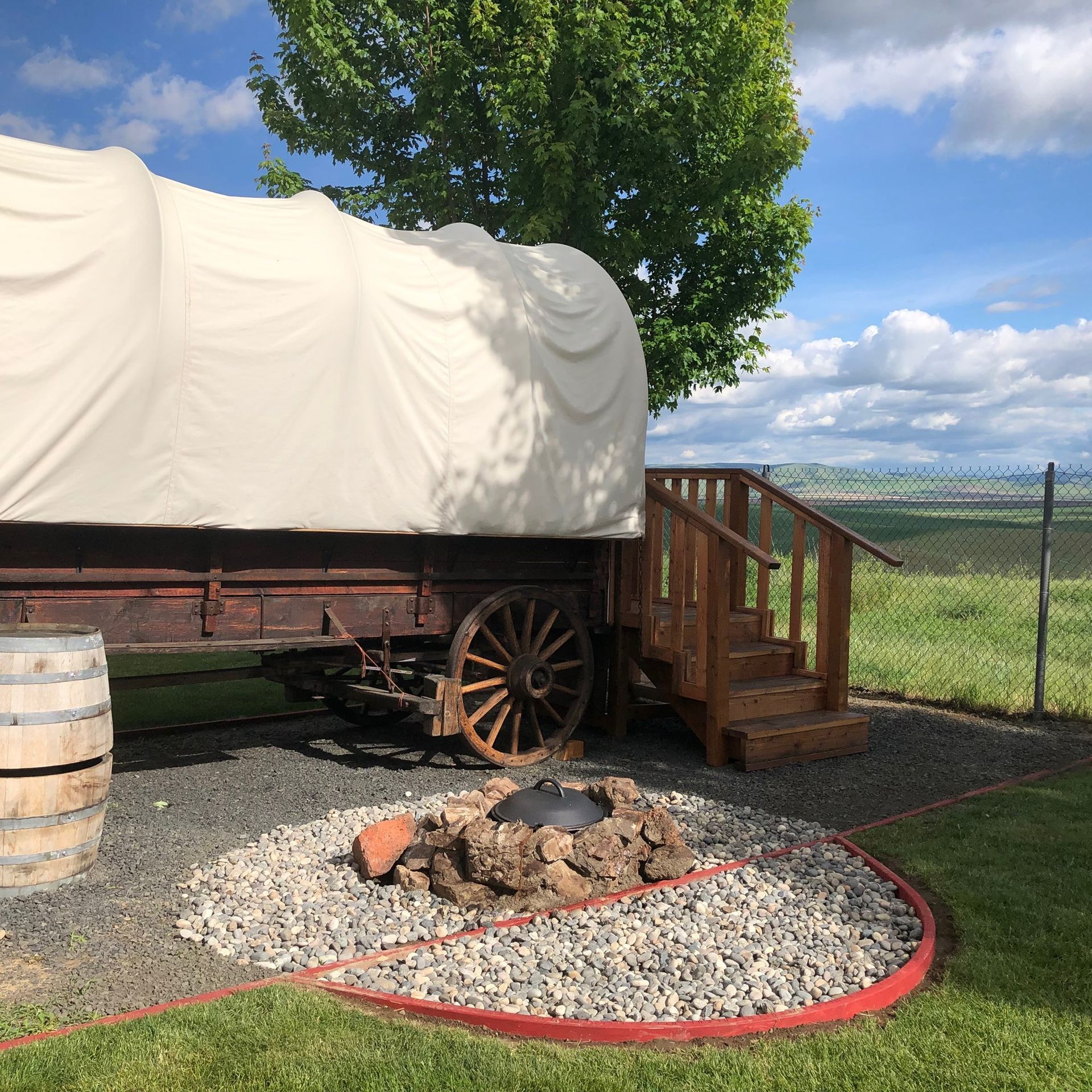 Covered wagon at the Pendleton KOA by a fire pit and barrel, with wooden steps and a fenced grassy field under a cloudy sky.