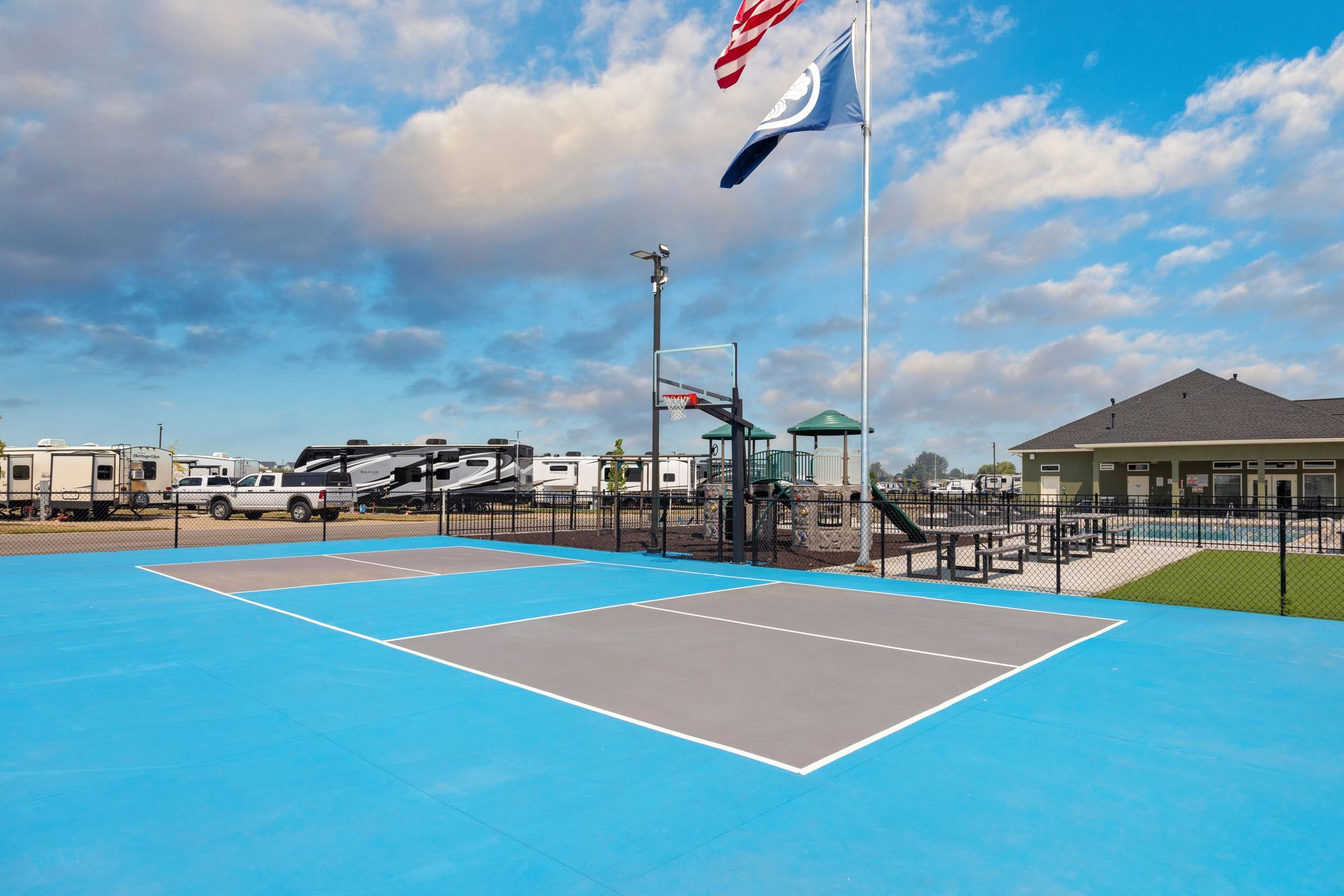 IdaHome RV Resort's outdoor basketball court with a blue and gray playing surface, flagpoles, and a cloudy sky.