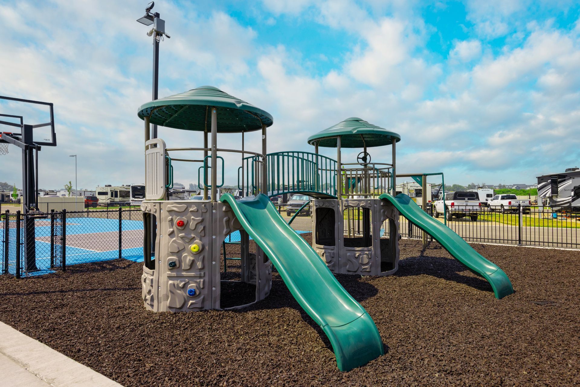 Playground equipment at Idahome RV Resort with two green slides and climbing structures on wood chips under a blue sky.