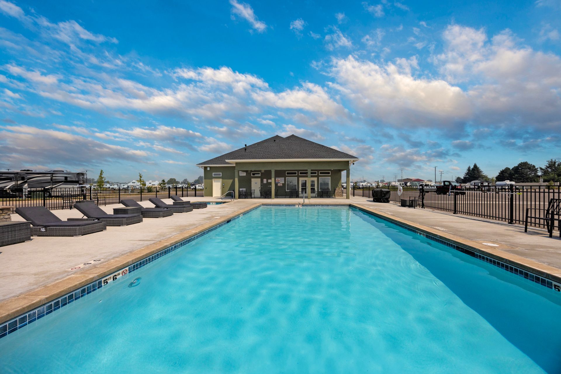 Outdoor swimming pool with lounge chairs and a cabana under a partly cloudy blue sky at IdaHome RV Resort.