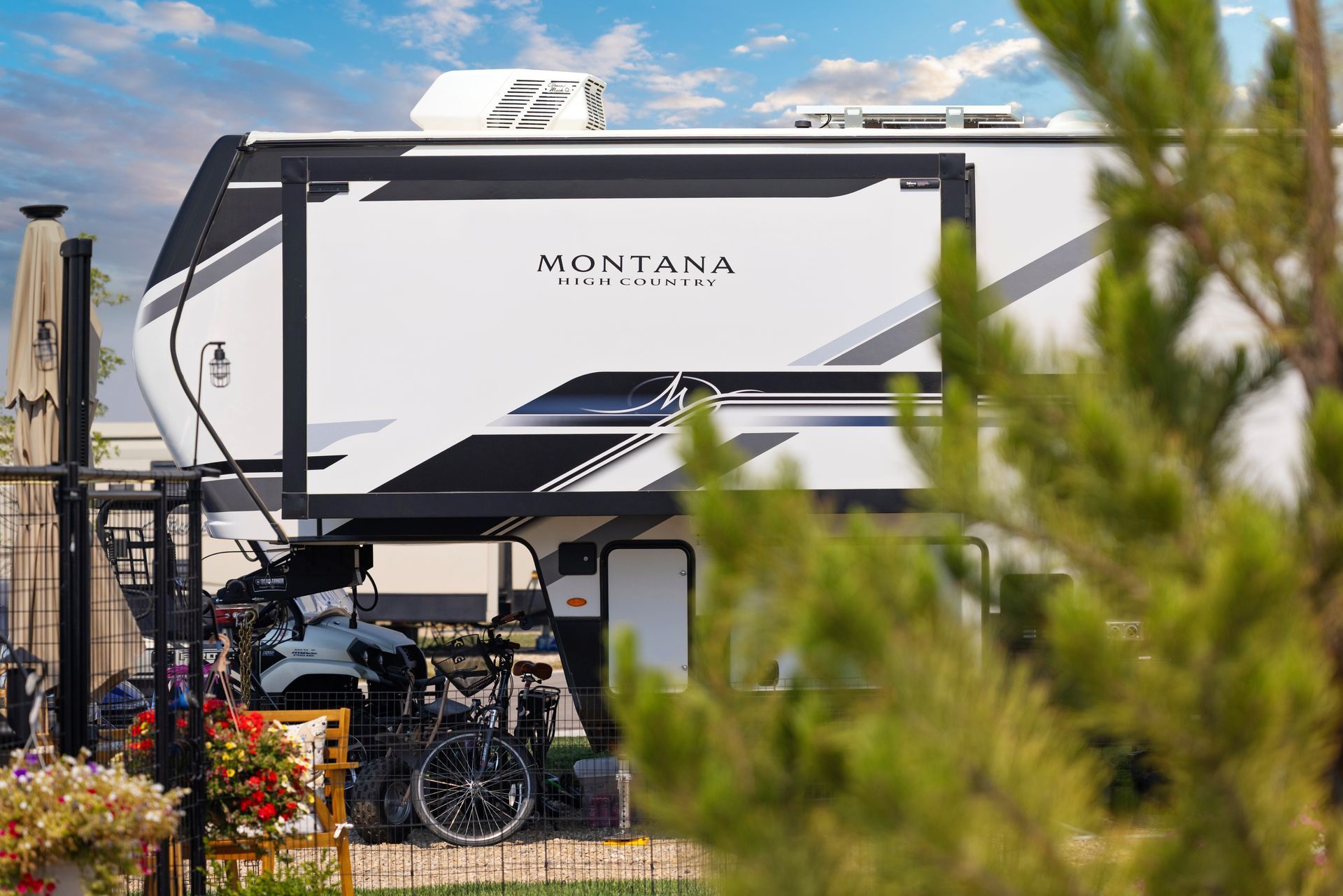 White Montana RV parked at the Idahome RV Resort with bicycles and camping gear, partly framed by trees and fence.