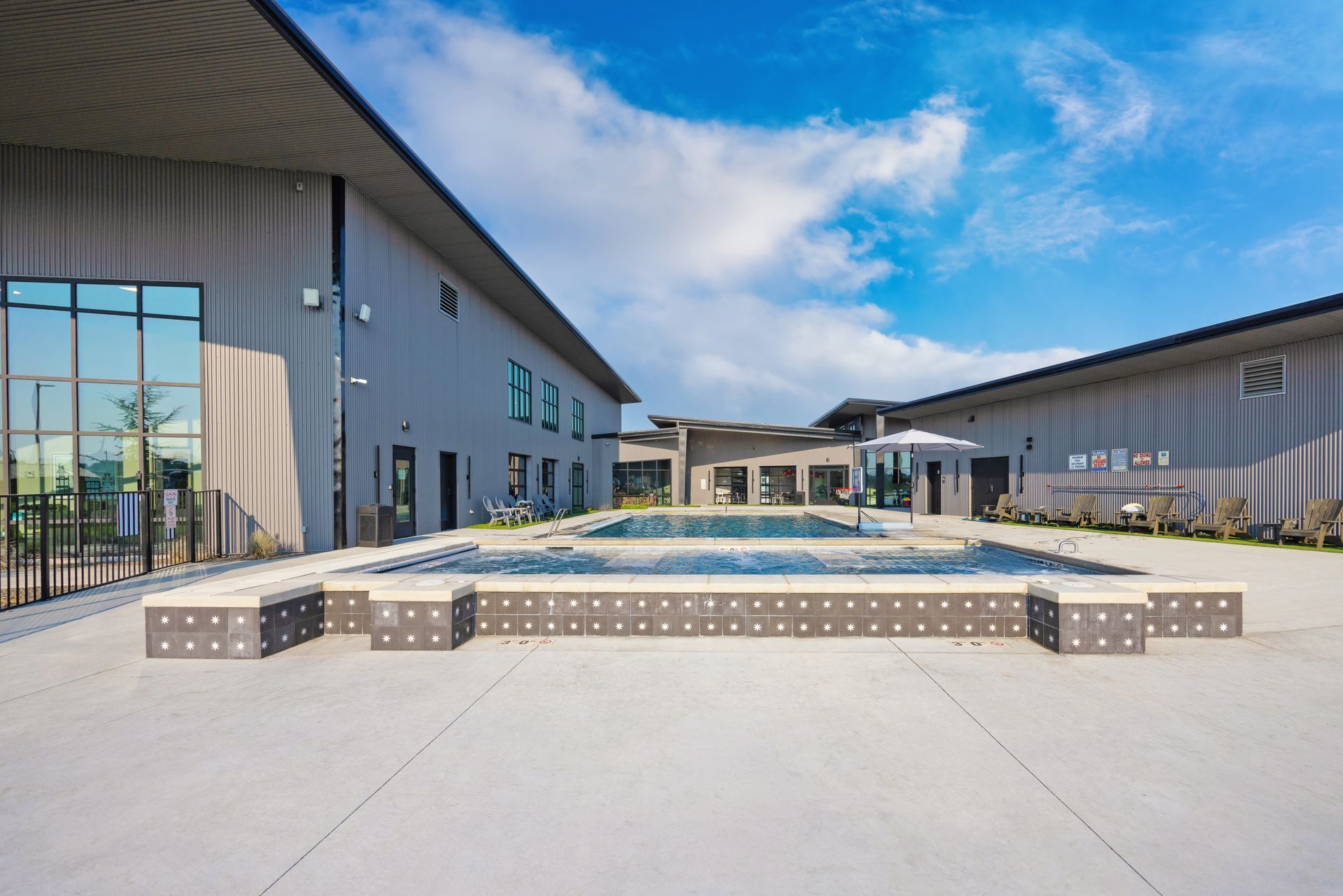 A modern outdoor pool and hot tub with raised brick seating, central water feature, and industrial-style buildings under a blue sky at the Sun Creek Point RV Park.