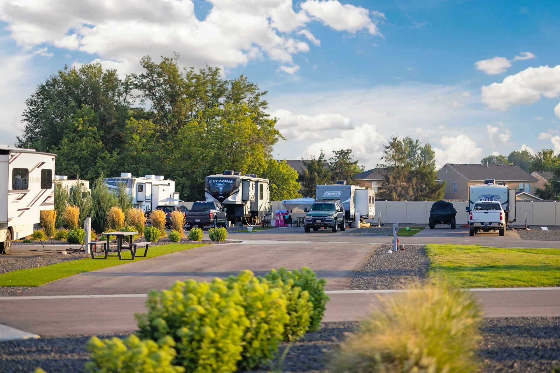 RVs and trailers parked in pull through sites at Sun Creek Point RV Resort beside trees under a partly cloudy sky.