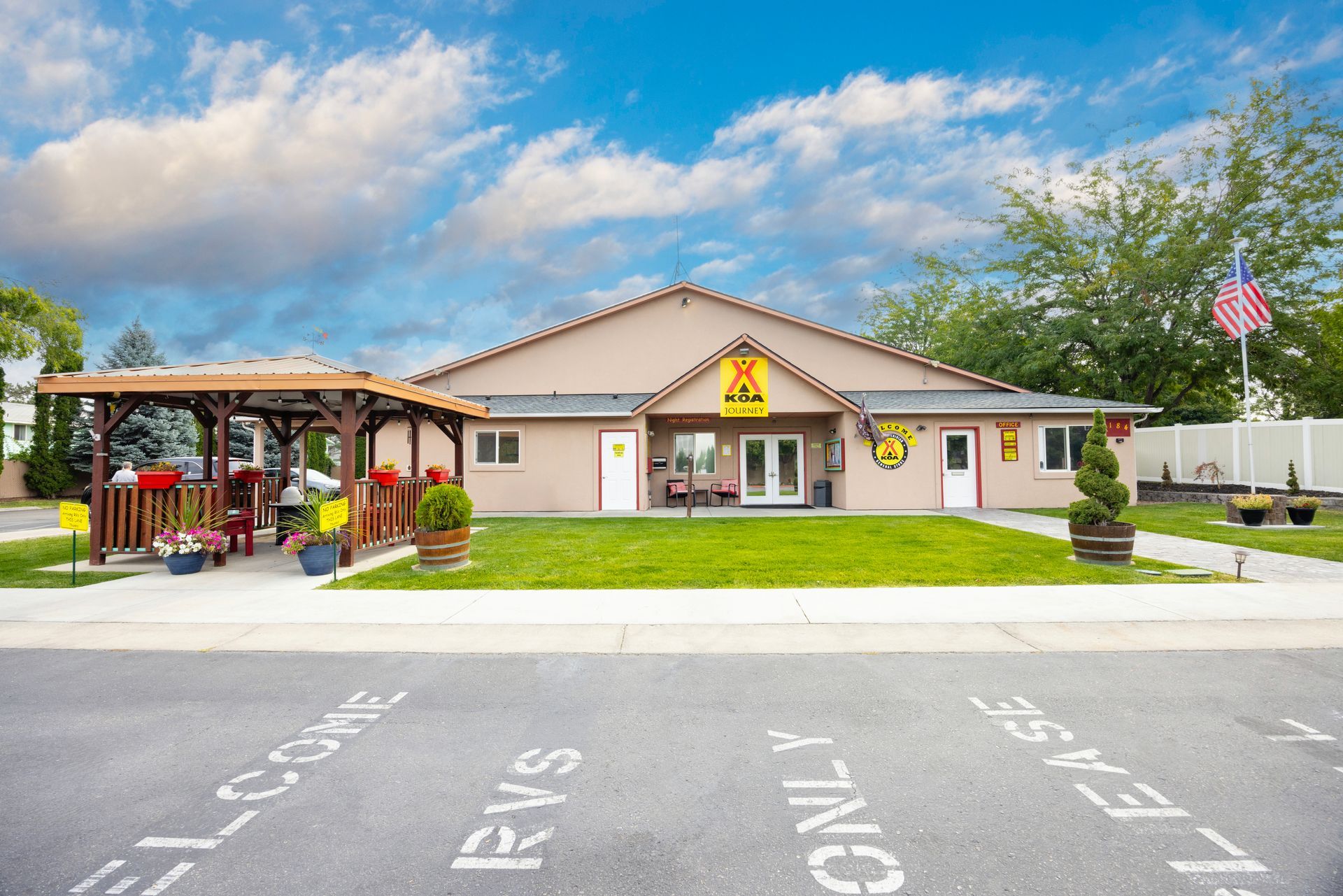 The Boise Meridian KOA club house with a covered patio, red flowers, grassy lawn, and American flag under a cloudy sky.