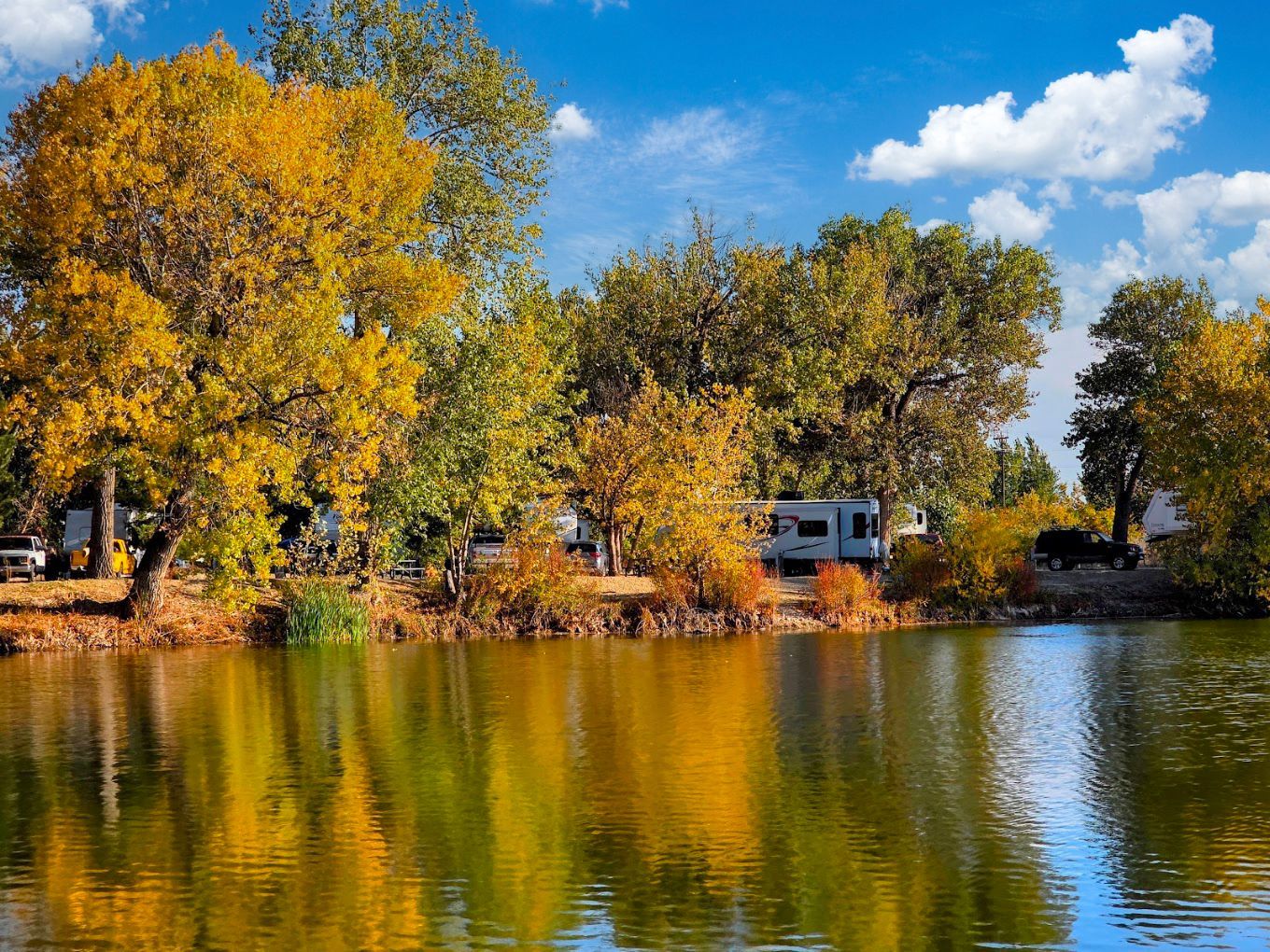 Autumn trees reflected in a calm lake under a blue sky with white clouds at Lake Point Campground.