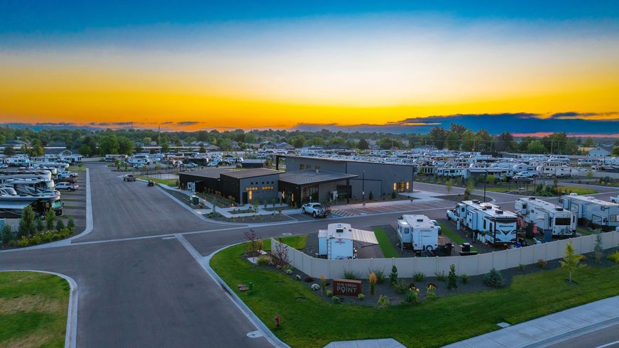 Sunset over a wide road and industrial site with trucks, buildings, and distant hills under a colorful sky.
