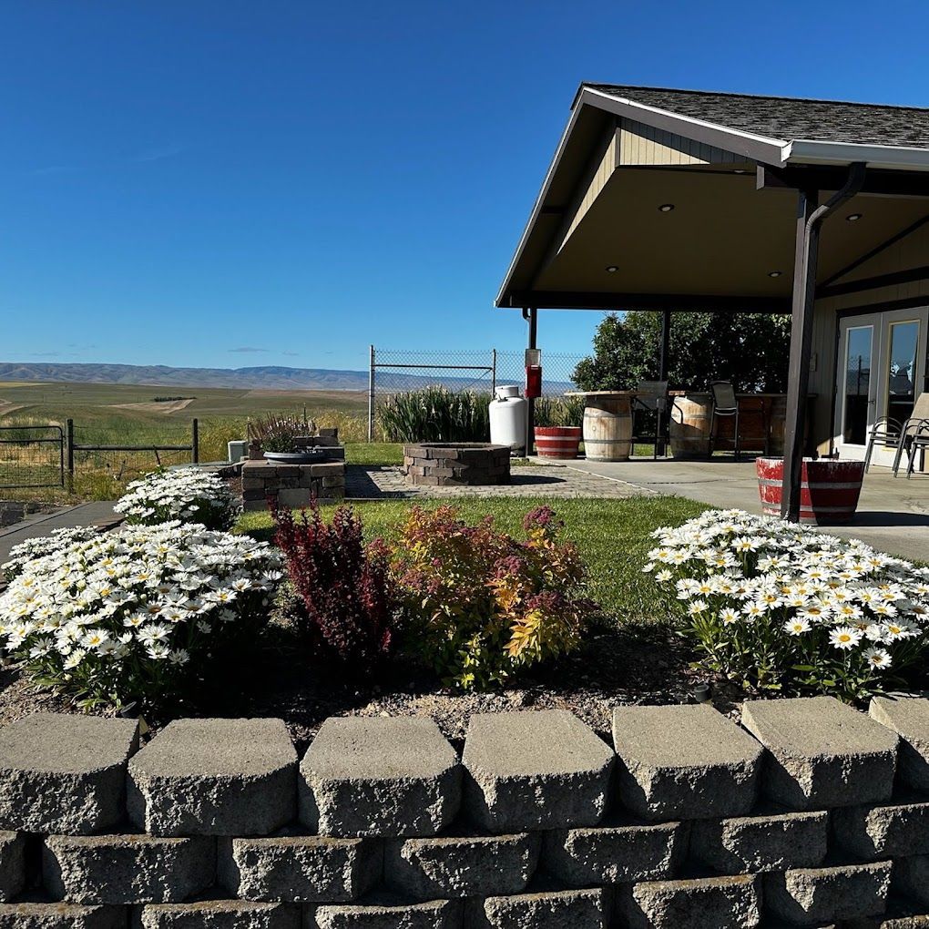 Pendleton KOA club house with with white flowers, potted plants, and a covered porch overlooking open fields under a clear blue sky.