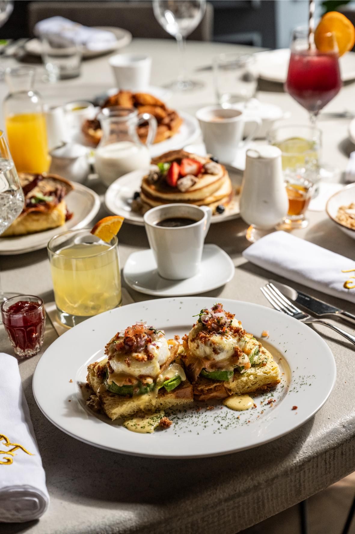 Disposición de la mesa para el brunch: huevos Benedict, panqueques, bebidas y café en una mesa de color claro.