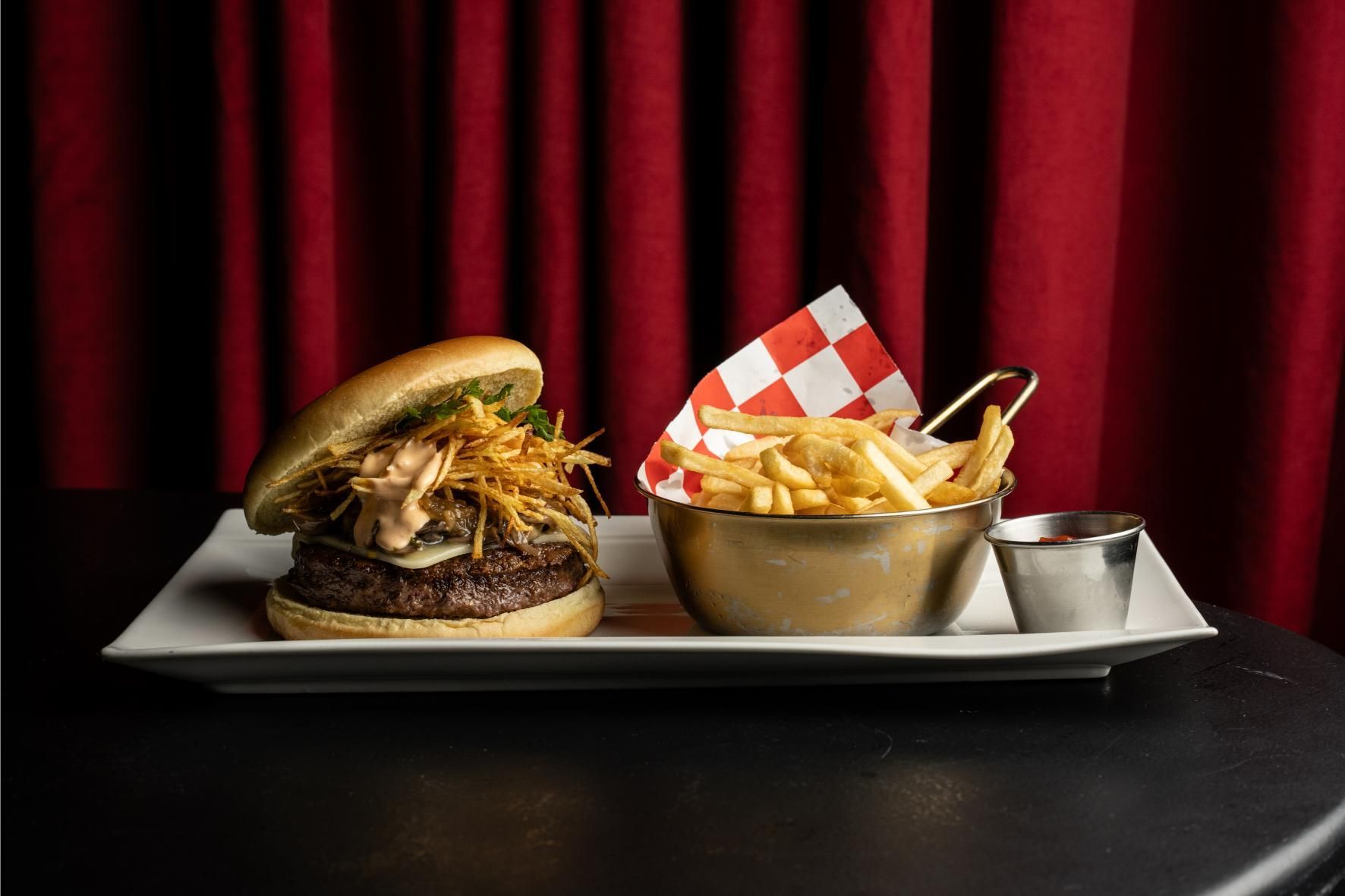 Hamburguesa con papas fritas en plato blanco, recipiente dorado, servilleta a cuadros rojos y blancos, fondo de cortina roja.