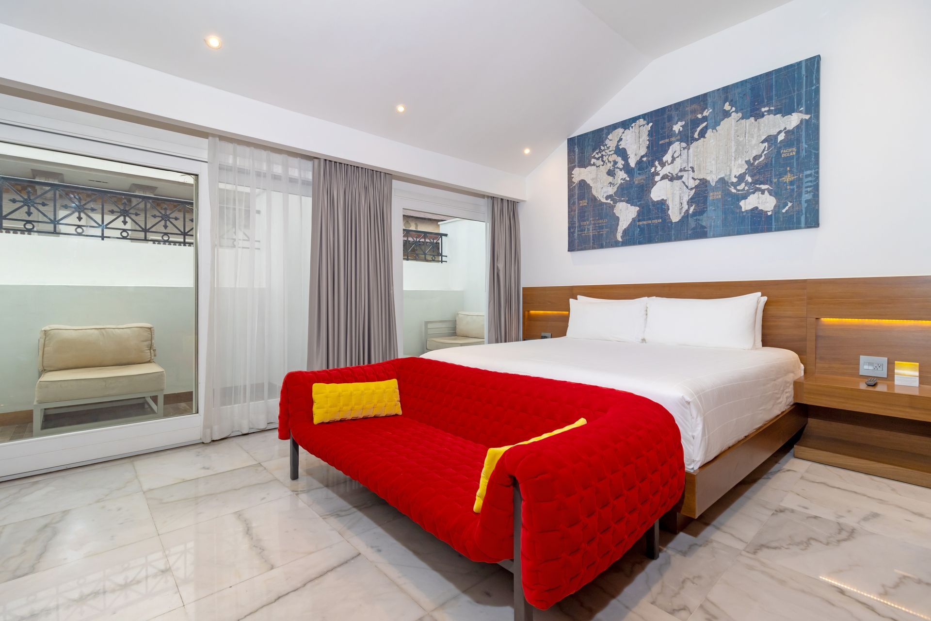 Dormitorio de hotel luminoso con sofá rojo, mapas artísticos y acceso al balcón.
