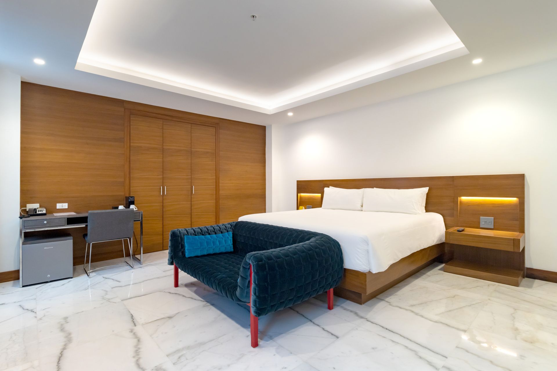 Dormitorio de hotel con pisos de mármol, cama King, sofá azul, armario de madera y escritorio.