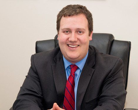 Smiling man in a dark suit and red tie seated at a desk in an office chair