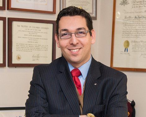 Smiling man in a dark suit and red tie, seated in an office with framed certificates on the wall