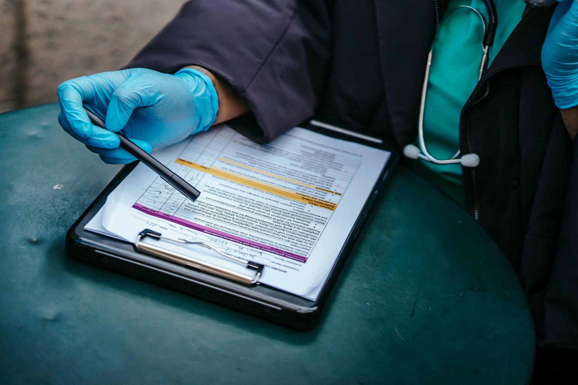Gloved clinician points to a clipboard checklist on a table with a stethoscope nearby.