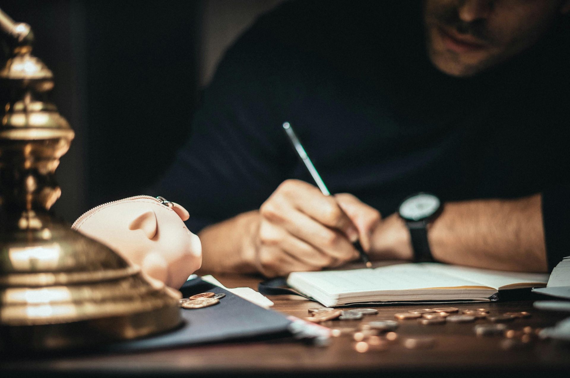 Person writing in a notebook beside a warm-lit desk lamp and a white mask on a dark table