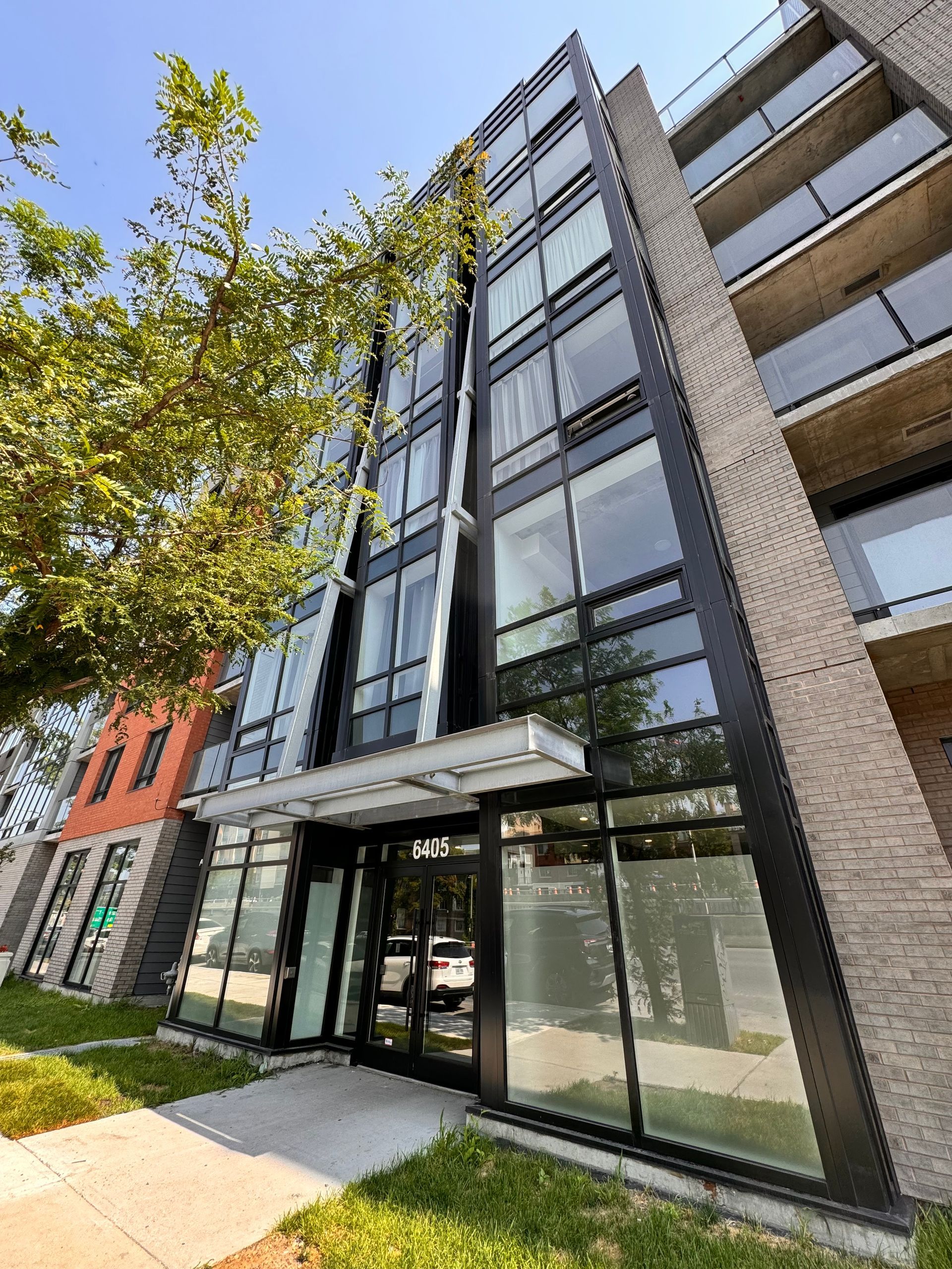 Bâtiment moderne avec entrée et fenêtres vitrées, façade en briques et métal sombre. Arbre vert devant.