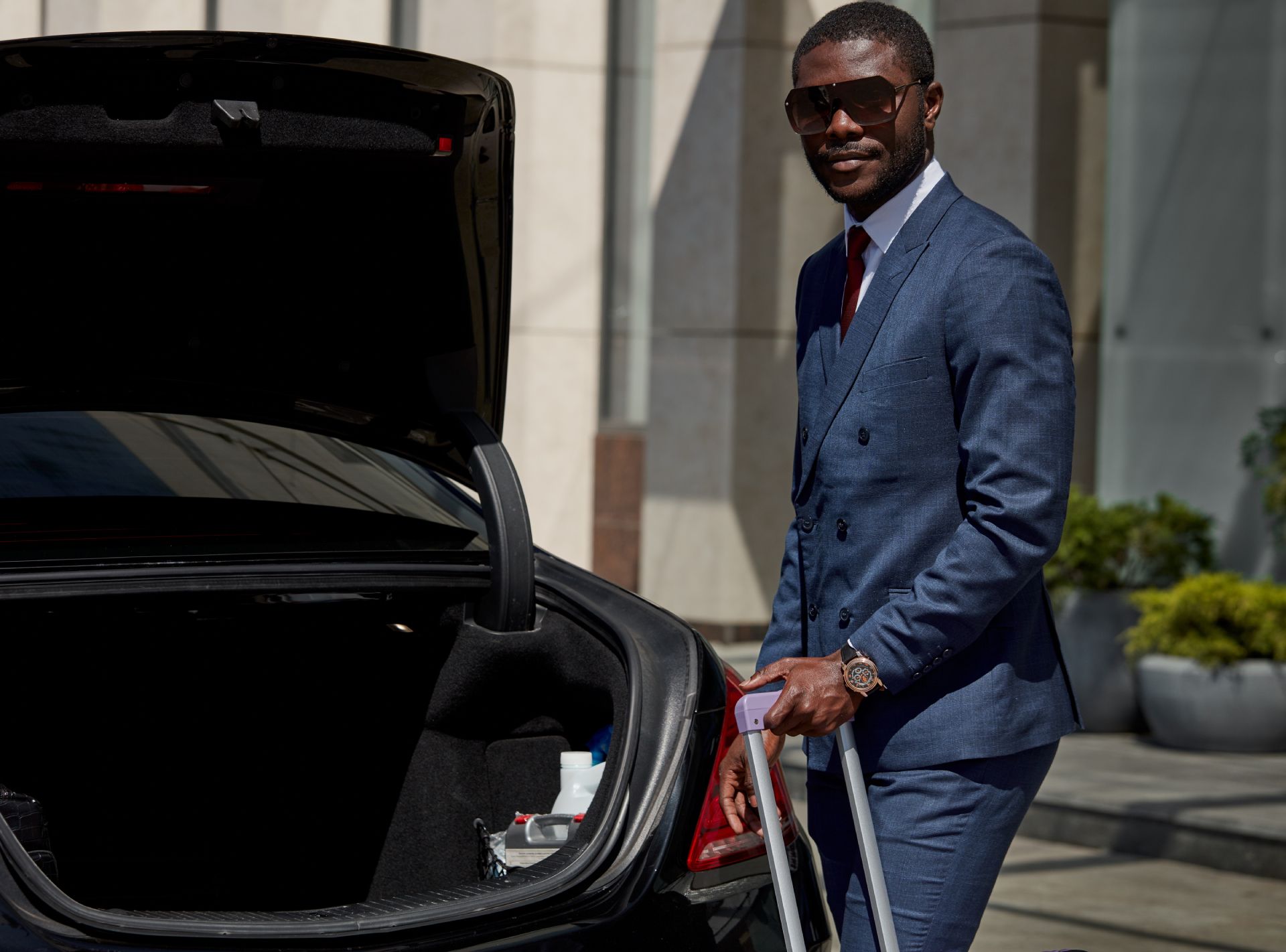 Man standing by an open car trunk in a suit and sunglasses, holding luggage outside a building.