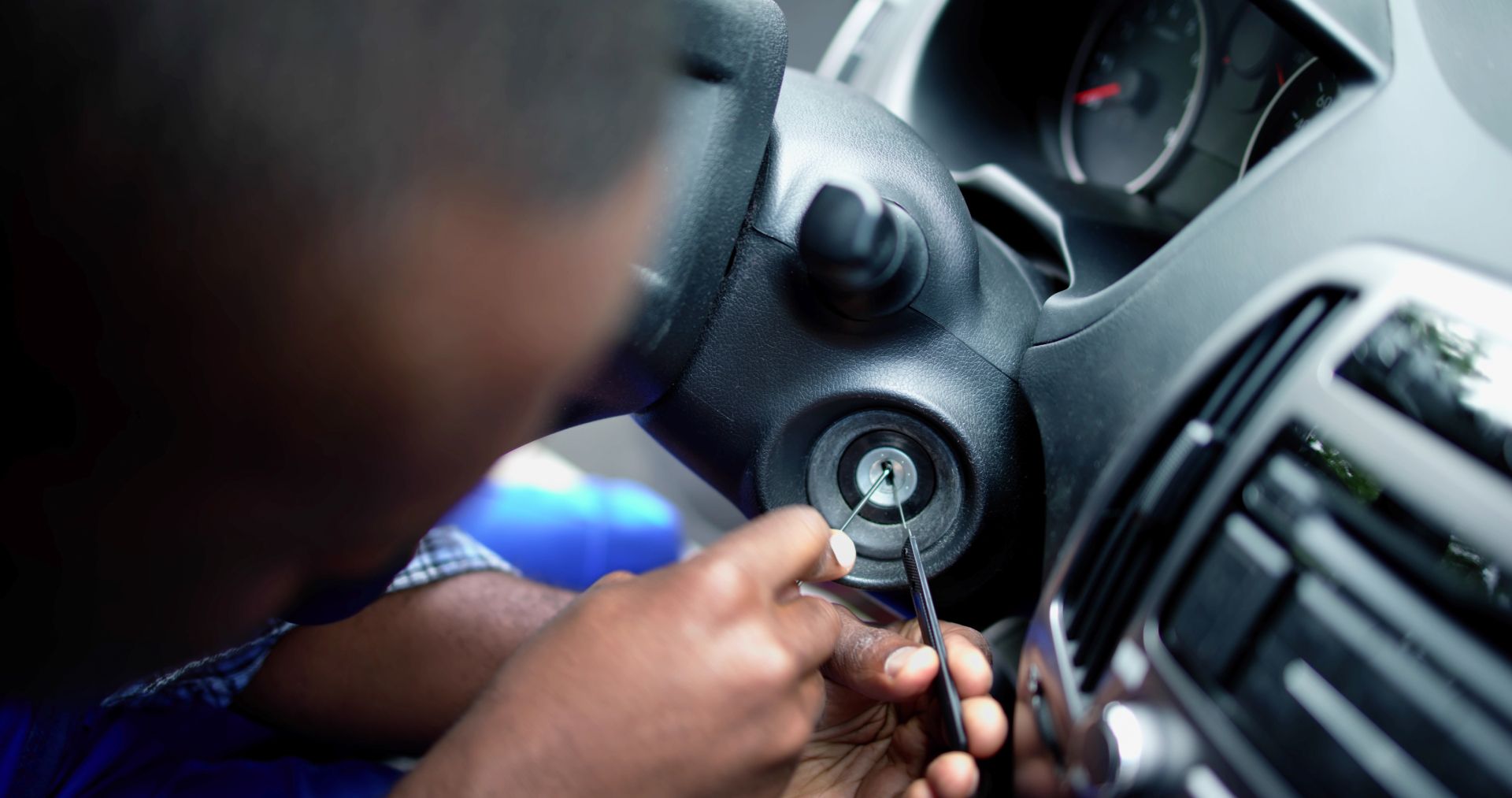 Hands turning a car ignition key on the steering column beside the dashboard.
