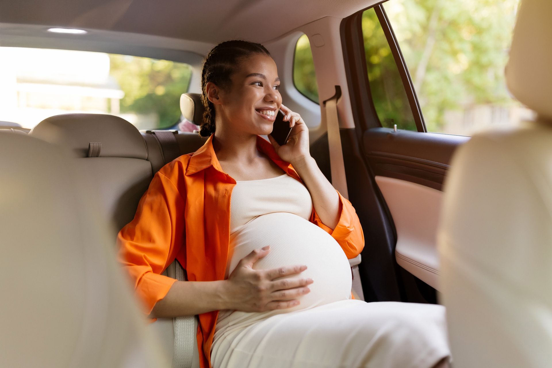 Pregnant woman in an orange shirt sits in a car, smiling while talking on a phone.