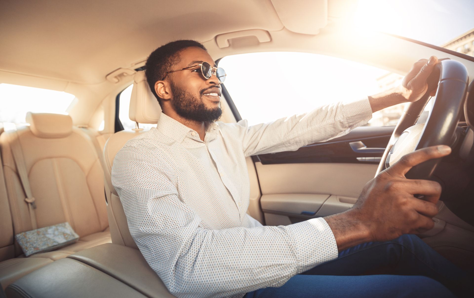 Man driving a car, smiling in a sunlit beige interior while holding the steering wheel