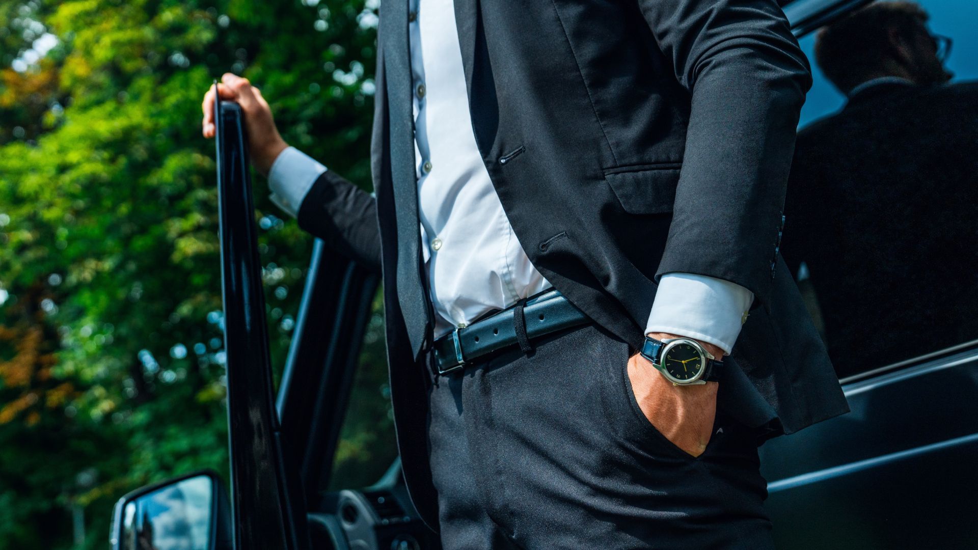 Person in a black suit stands by an open car door, wearing a white shirt and watch.