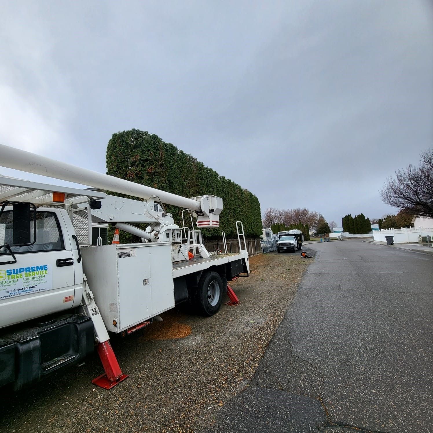 White utility truck with crane parked on a gravel roadside under cloudy sky, near trimmed hedges.