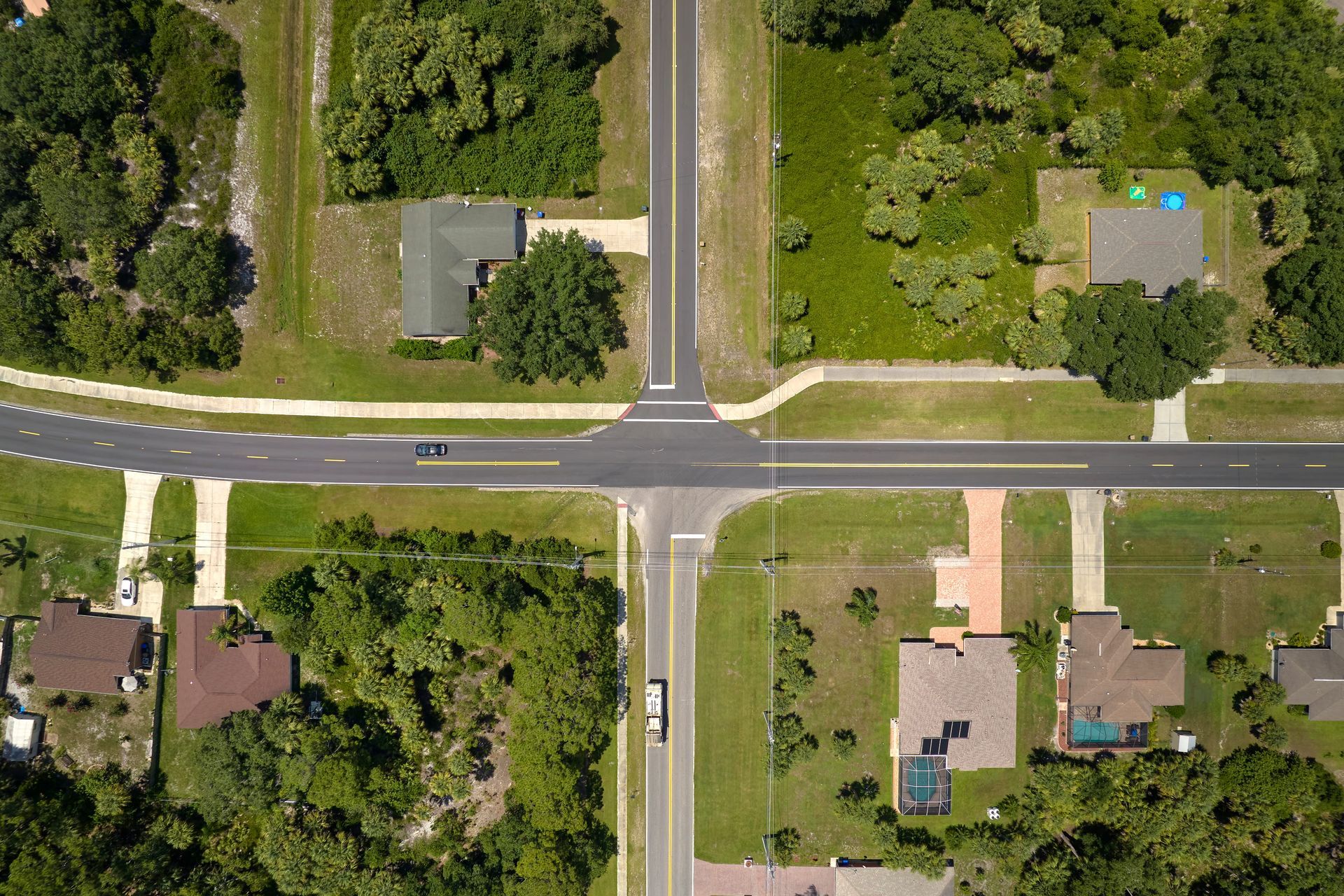 Aerial view of a suburban four-way intersection with houses, trees, lawns, and parked cars.