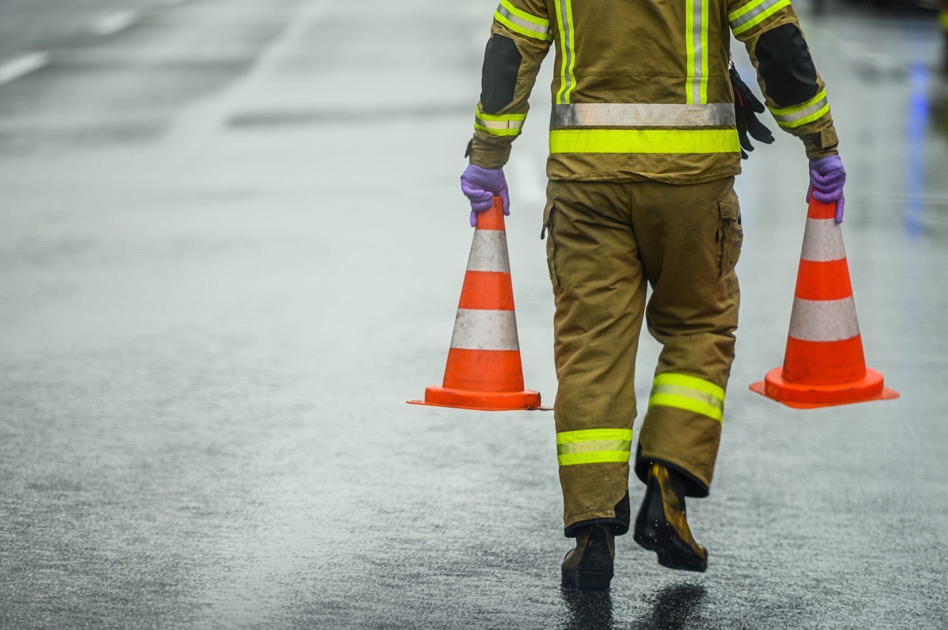 Road worker walking on wet pavement carrying orange traffic cones, reflective safety gear visible