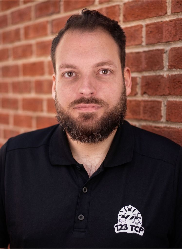 Adam from 123TCP in a black polo shirt standing before a red brick wall, looking at the camera.