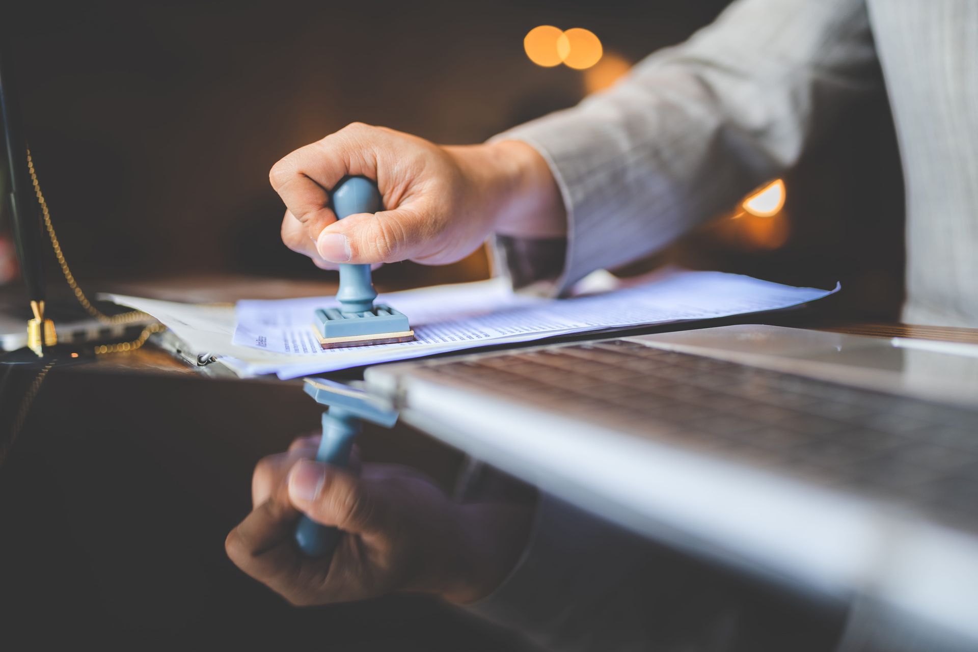 Hand stamping a document on a desk, with a blurred laptop in the background