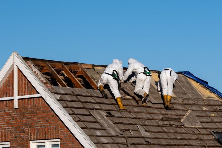 Three workers in white suits repair a damaged roof on a brick house under a blue sky.