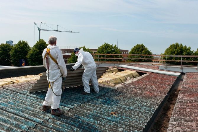 Two workers in white protective suits inspect a rooftop covered with a blue and red mesh tarp.