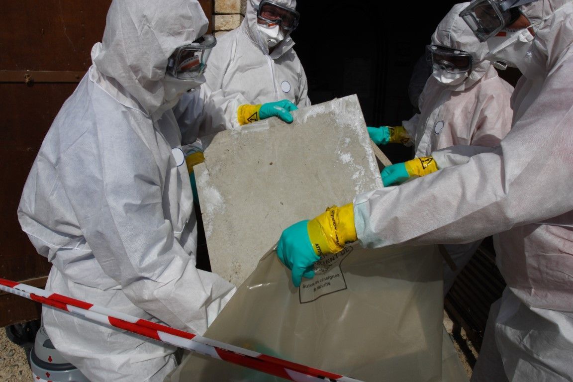 Workers in white hazmat suits handle a large block of material at an outdoor site.