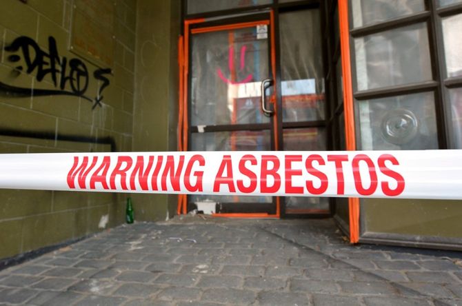Red-and-white “WARNING ASBESTOS” tape blocking a building entrance with broken windows and graffiti