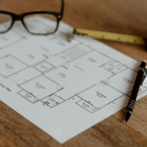 Architectural floor plan on a wooden desk with eyeglasses, pen, and tape measure.