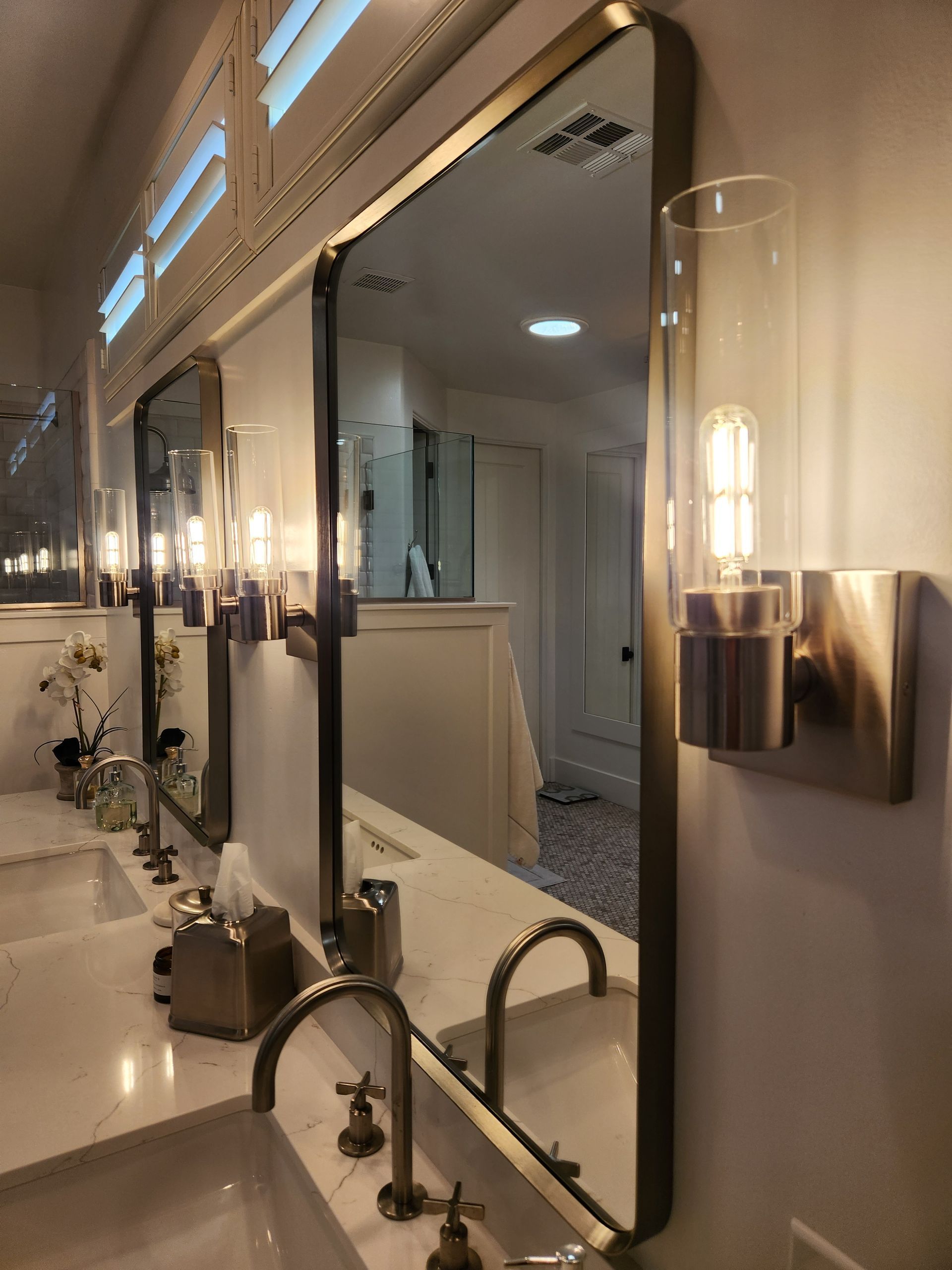 Bathroom vanity with mirrors and lit wall sconces along a sink counter.