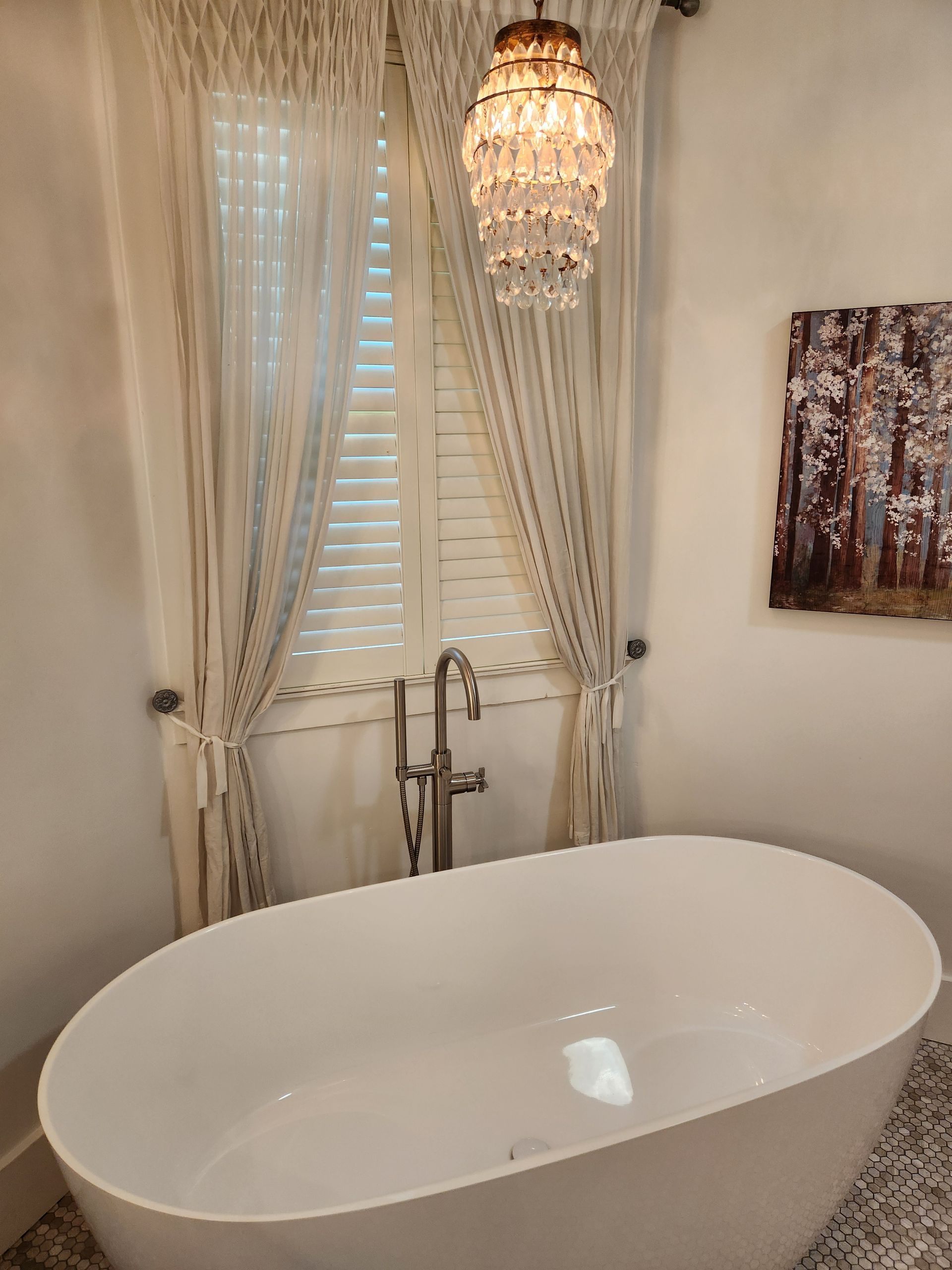 Elegant bathroom with a white oval tub, chandelier, and sheer curtains over a window.