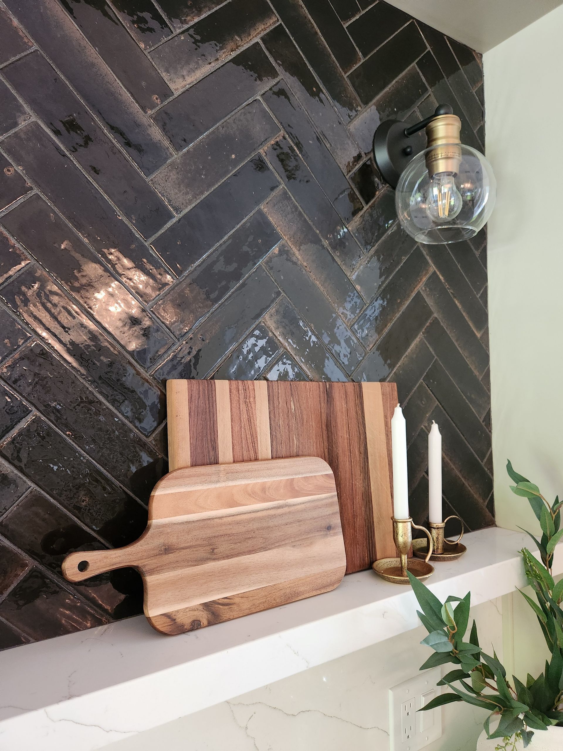 Kitchen shelf with herringbone tile backsplash, wooden cutting boards, candles, and a glass light fixture.