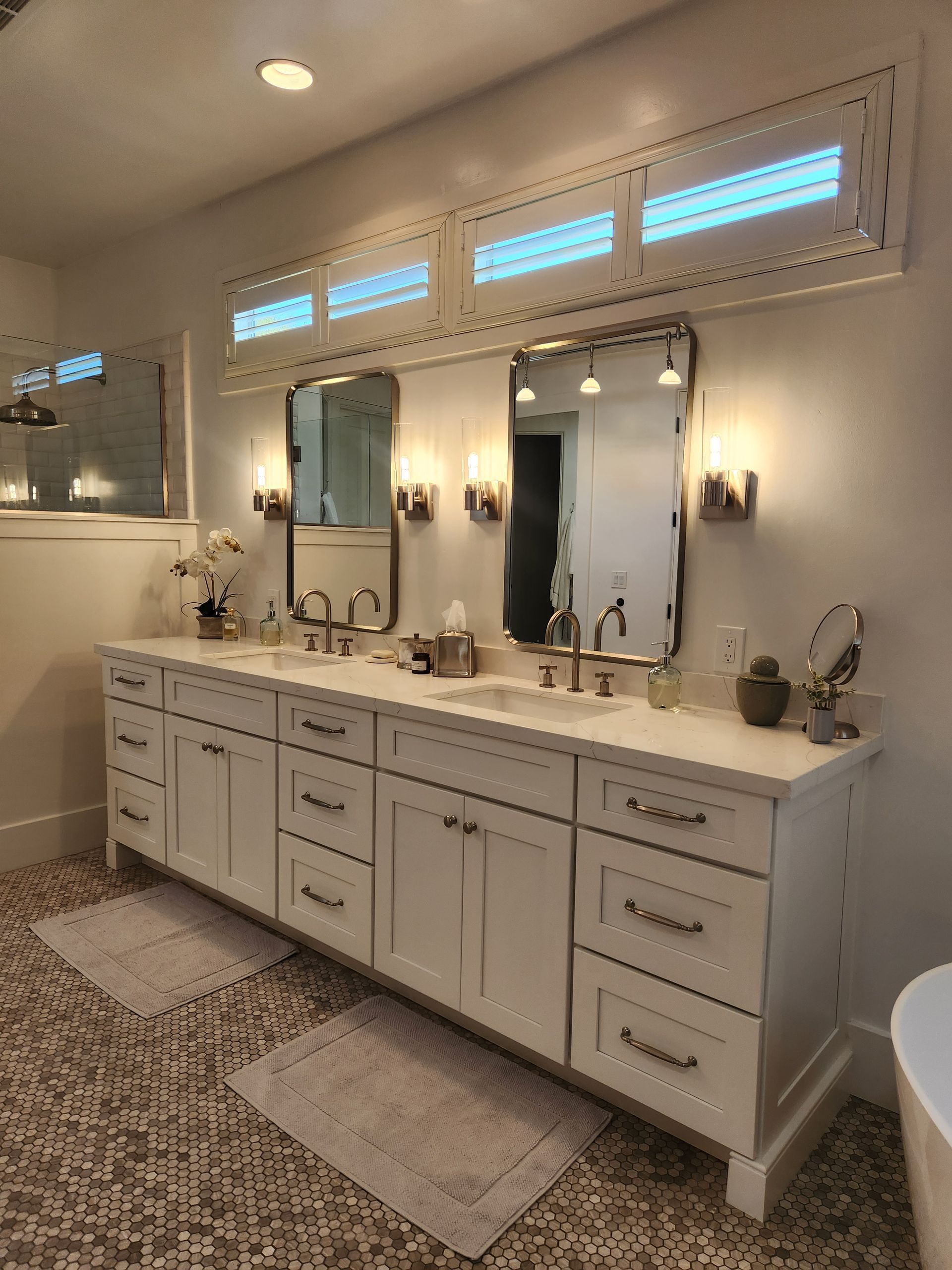 Elegant bathroom vanity with double sinks, mirrors, sconces, and patterned rugs under high windows