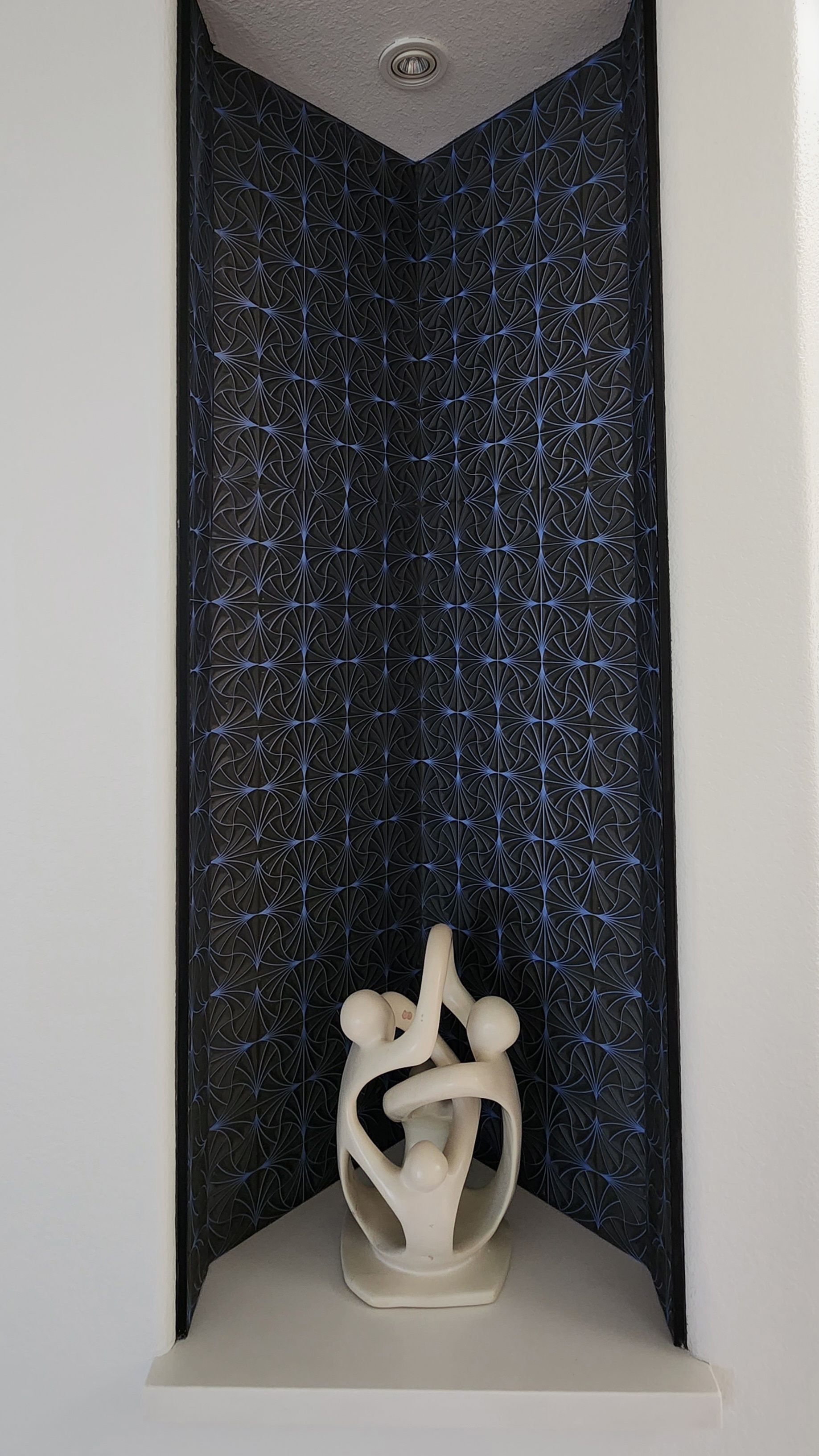 Abstract white sculpture in a recessed wall niche with dark patterned backing and a ceiling light.